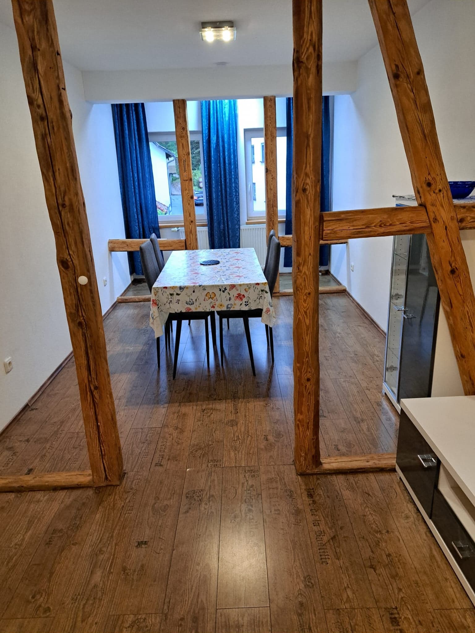 Aufenthaltsraum der Wohnung A mit freiliegenden Fachwerkbalken, Esstisch für vier Personen und blauen Vorhängen